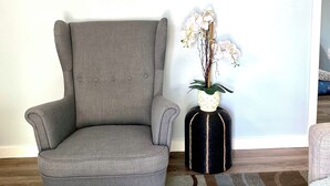 Un sillón gris, un espejo redondo y un florero con flores blancas frente a una pared azul claro.