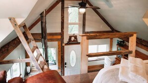 A loft bedroom with a sloped ceiling, wooden beams, a ceiling fan, and a view of the outside. - Relax at this cozy, well appointed cabin located in sunny Knik, Alaska.  (Wasilla)