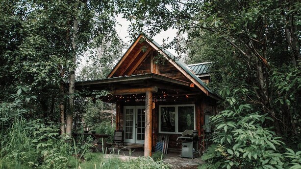 A rustic cabin with a porch, surrounded by dense forest. - Relax at this cozy, well appointed cabin located in sunny Knik, Alaska.  (Wasilla)