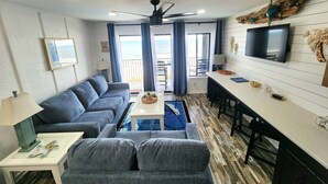 A modern living room with a sofa, coffee table, and TV. There is a balcony with a view of the sea.