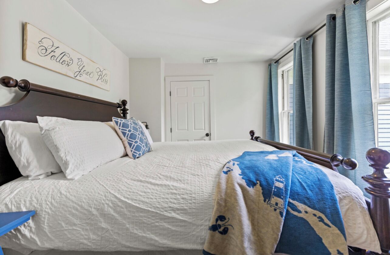 A neatly made bed with a white comforter and a blue and white blanket, a wooden headboard, and a wall with a sign that reads "Follow Your Bliss".