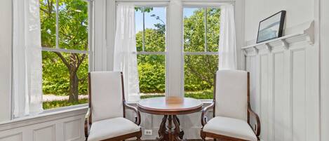 A room with a round wooden table and two chairs, large windows with white curtains, and a view of greenery outside.