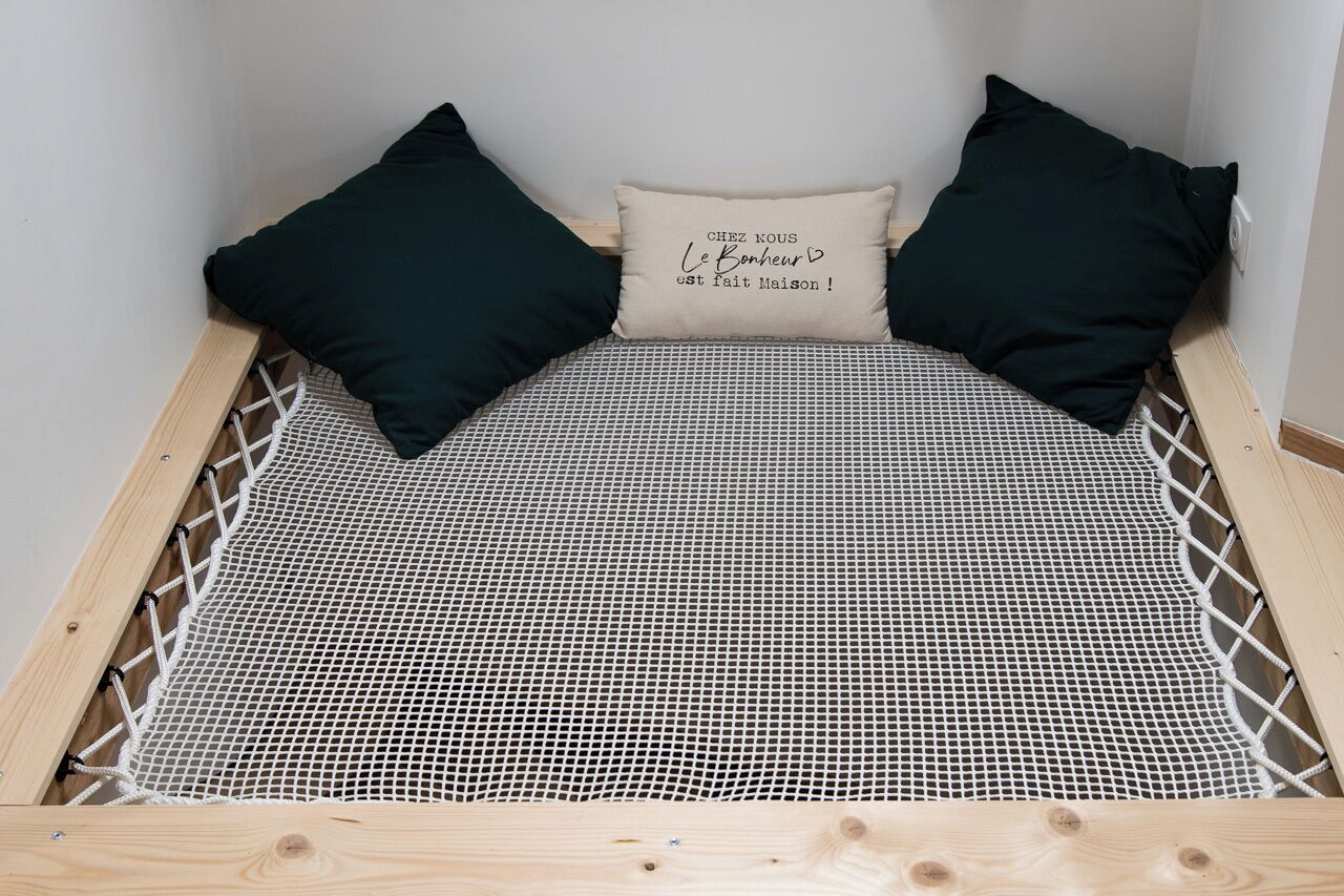 Een bed met een witte netdeken, twee donkere kussens en een beige kussen met Franse tekst.