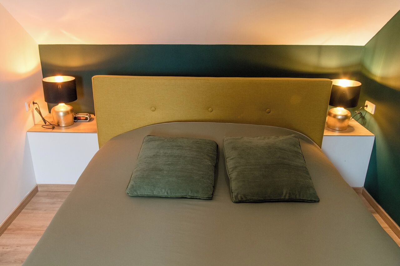 Een bed met twee kussens, een hoofdbord, nachtlampjes en een klein tafeltje met een lamp en een klok.