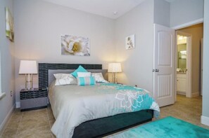 A bedroom with a bed, two bedside tables, a nightstand, a door, and a bathroom visible through an open door. - Sunshine at Solterra (Davenport)