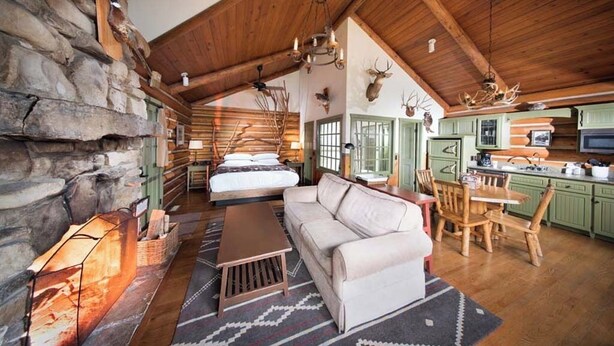 Interior de una cabaña de troncos con chimenea de piedra, una sala de estar acogedora con sofá y mesa de centro, un comedor con sillas de madera y una cocina con armarios verdes.