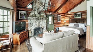 Interior de una cabaña de troncos con chimenea de piedra, televisor, sofá y cama.