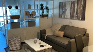 Una sala de estar moderna con un sofá gris, una mesa de centro blanca y dos otomanas tejidas. En el fondo se ve un estante blanco con adornos y plantas.