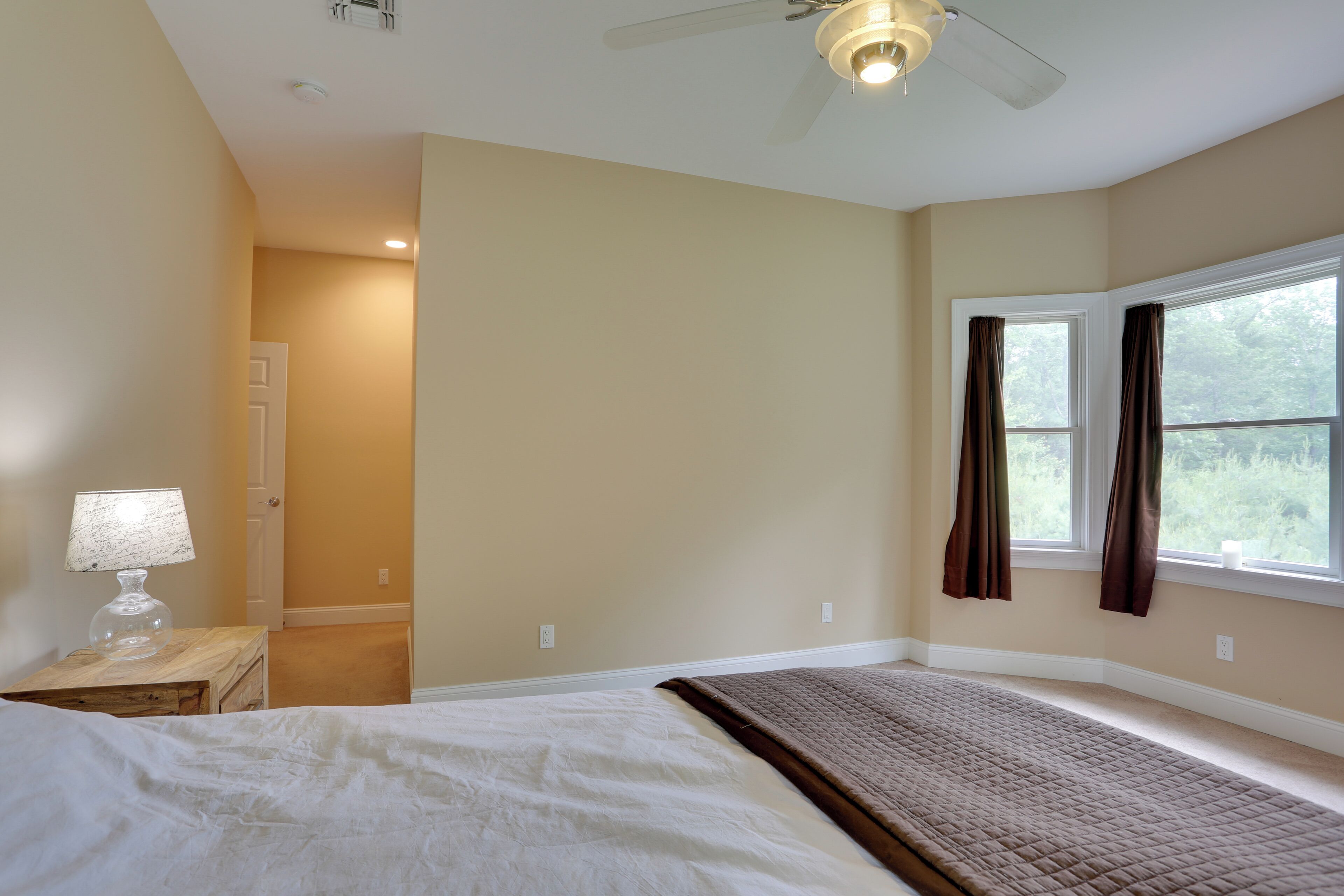 A bedroom with a bed, a nightstand with a lamp, a window with curtains, and a ceiling fan.