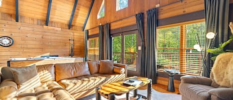 A spacious living area with a high ceiling, wooden walls, a sofa, a coffee table, and a view of greenery outside.