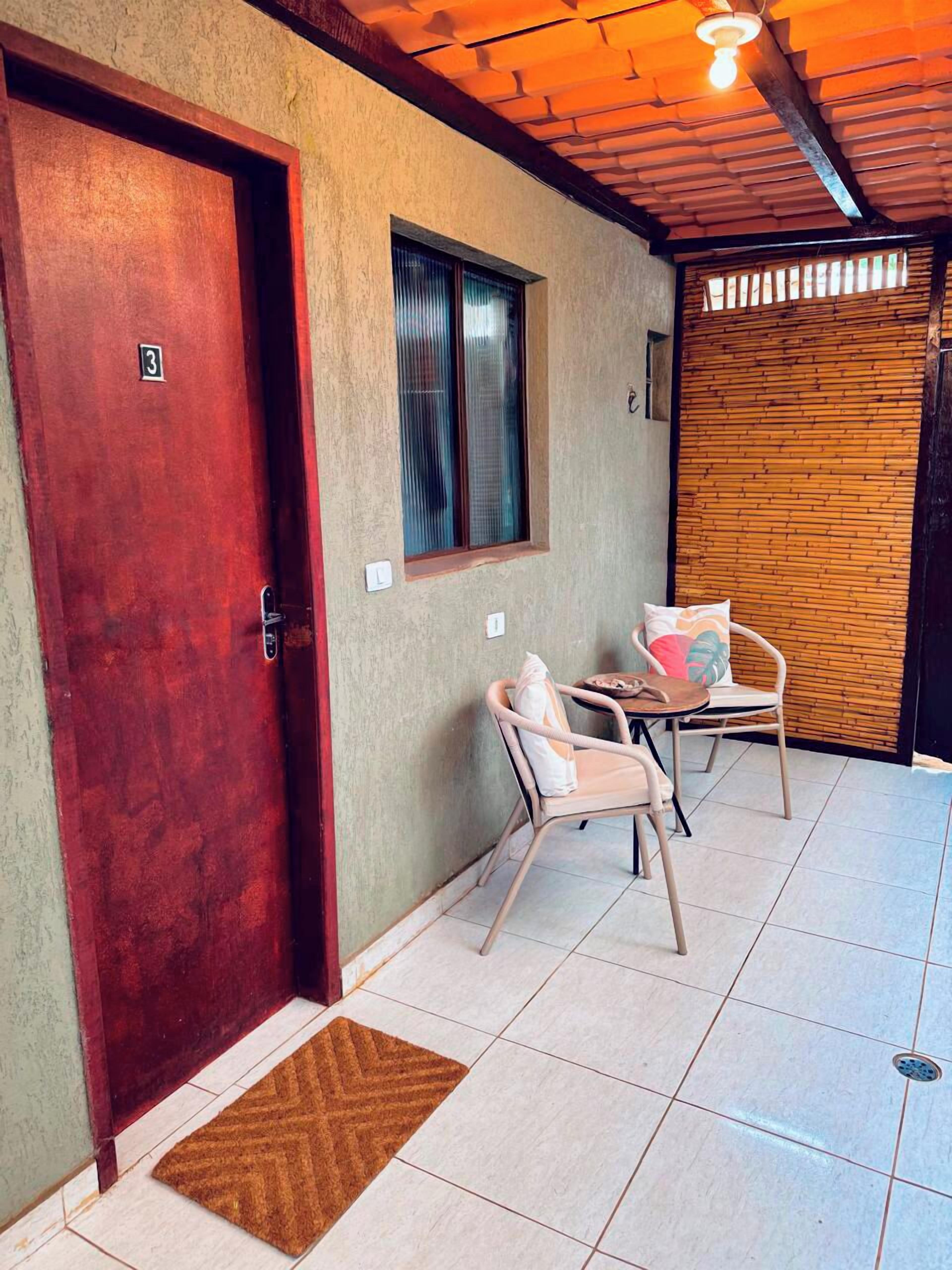 A room with a wooden door, a window, a wall-mounted light fixture, a doorbell, a small table with chairs, and a tiled floor.