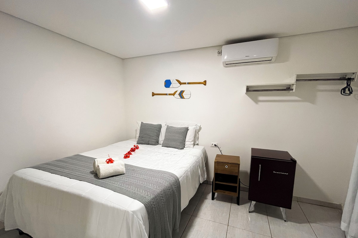 A hotel room with a bed, a nightstand, a dresser, an air conditioning unit, and a wall-mounted towel rack.
