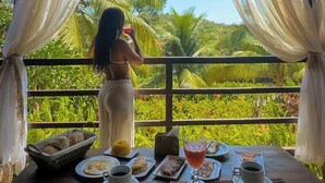 Uma área de refeições com vista para palmeiras, uma mesa posta com pratos, copos e uma cesta.