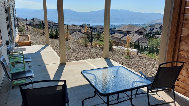Eine überdachte Terrasse mit Tisch und Stühlen, einer hölzernen Bank und Blick auf ein Wohngebiet mit Häusern und Bergen im Hintergrund.
