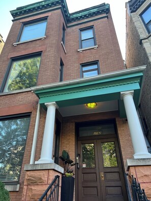 Un edificio de ladrillo con toldo verde, una puerta grande de color marrón y una placa con un número, además de un ventanal pequeño sobre la puerta.