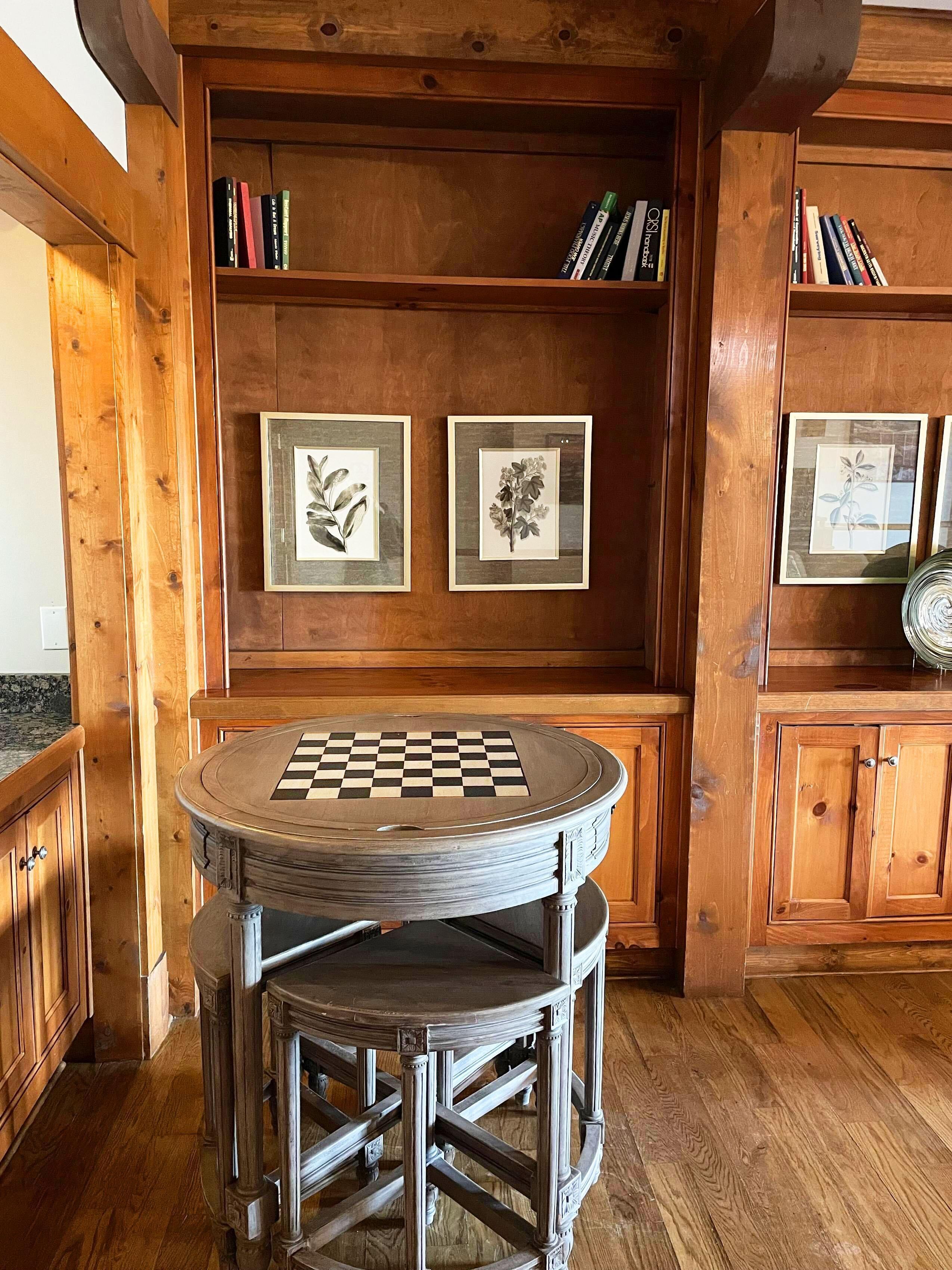 A wooden interior with a round table featuring a checkered surface, a framed botanical print, and a bookshelf with books.