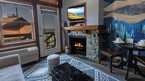 A cozy living room with a stone fireplace, a television mounted above it, and a large painting on the wall depicting a mountainous landscape. - HH 414 - Village/mountain views from this Charming Village studio! (Snowshoe)