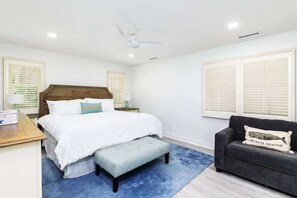 Une chambre à coucher comprenant un lit, un banc, une table de chevet, une commode, un canapé gris et un ventilateur de plafond.