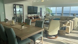 Un vaste espace de vie comprenant une table à manger en bois, des sièges confortables, un téléviseur et de grandes fenêtres offrant une vue sur l’océan.