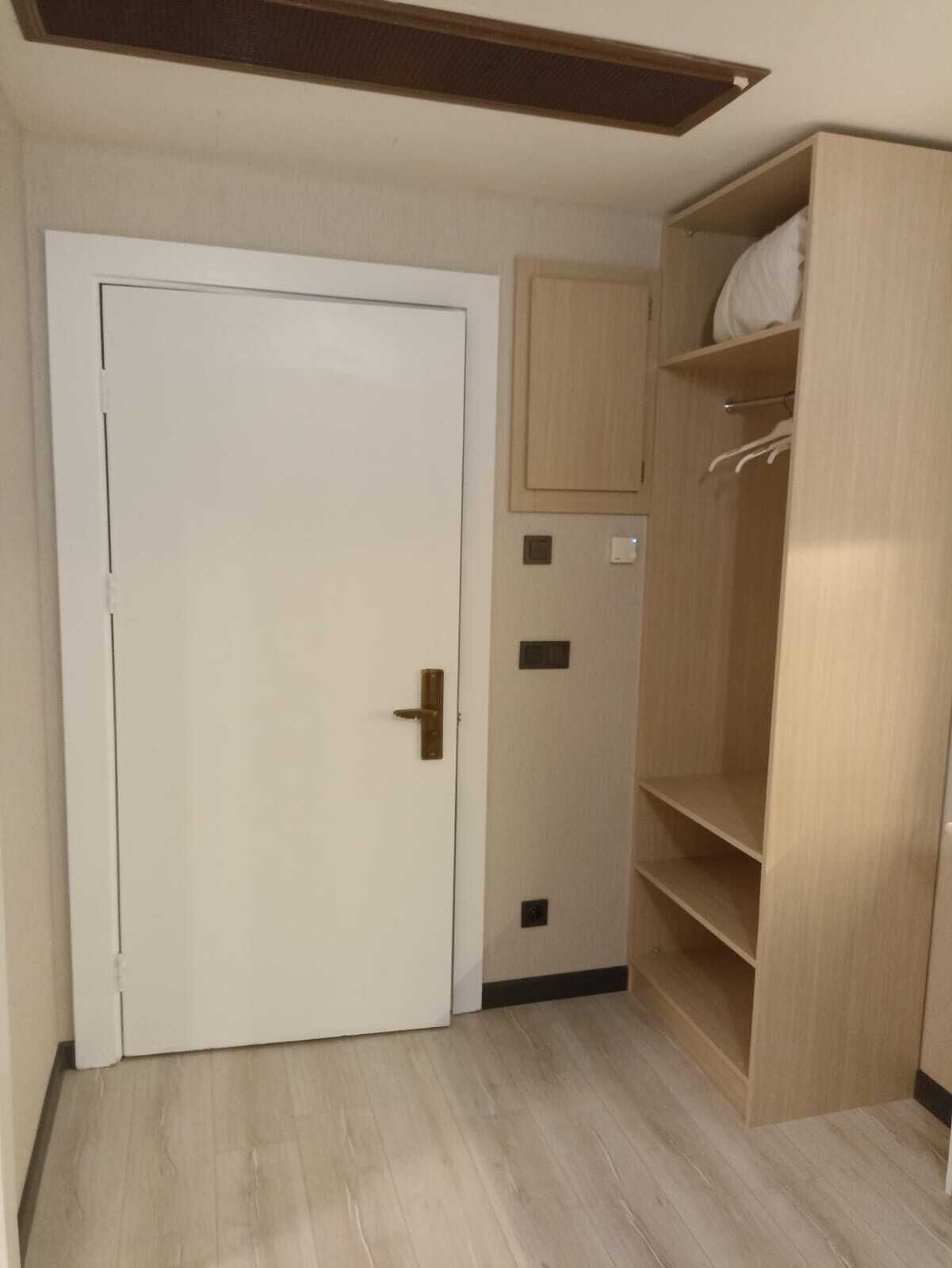 A room with a white door, a wooden wardrobe with shelves, and a small cabinet.