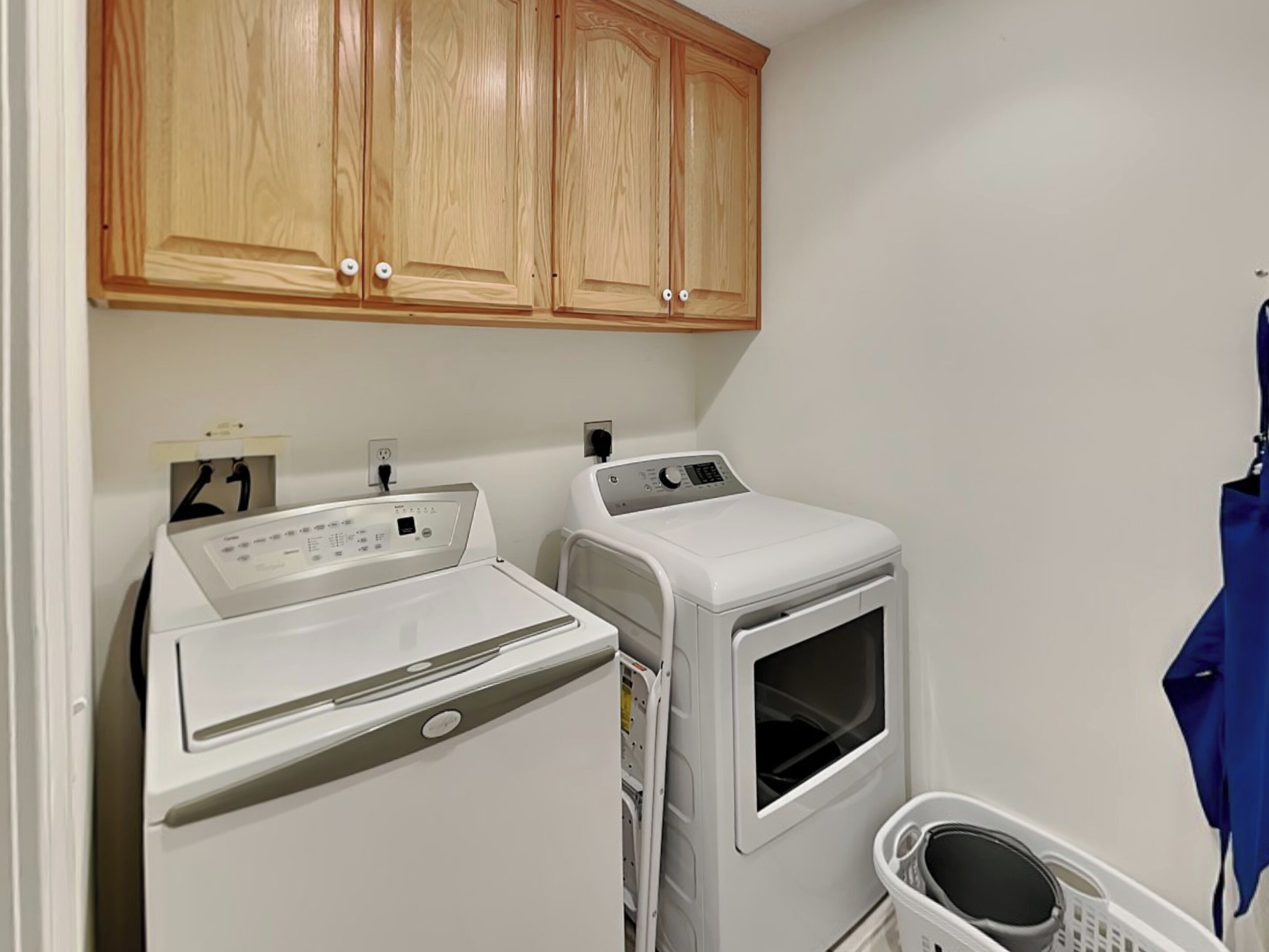 A laundry area with a front-loading washer and dryer, wooden cabinets above, and a laundry basket.