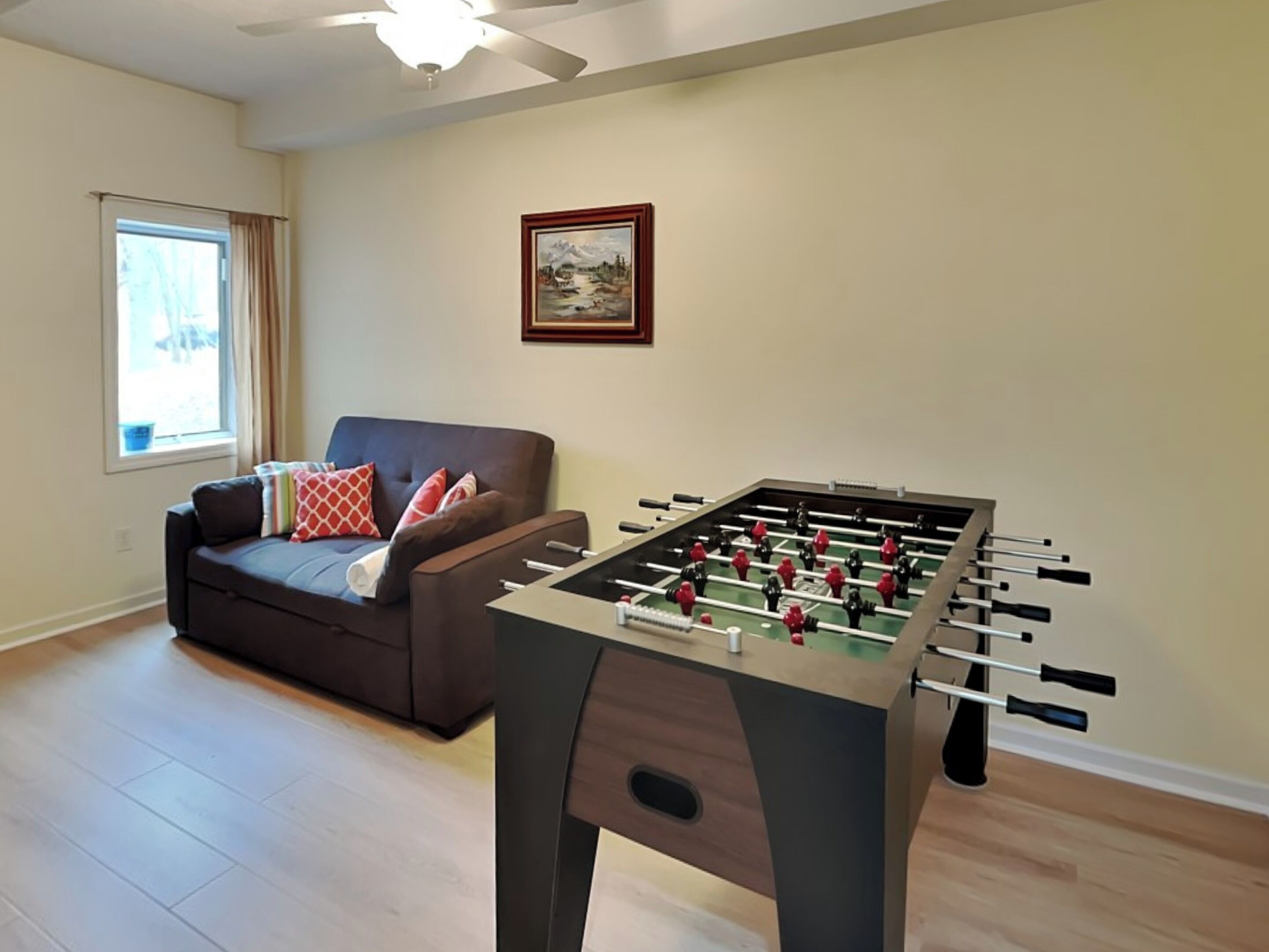 Un salon avec une table de baby-foot, un canapé avec des coussins, une fenêtre avec des rideaux et un tableau encadré au mur.