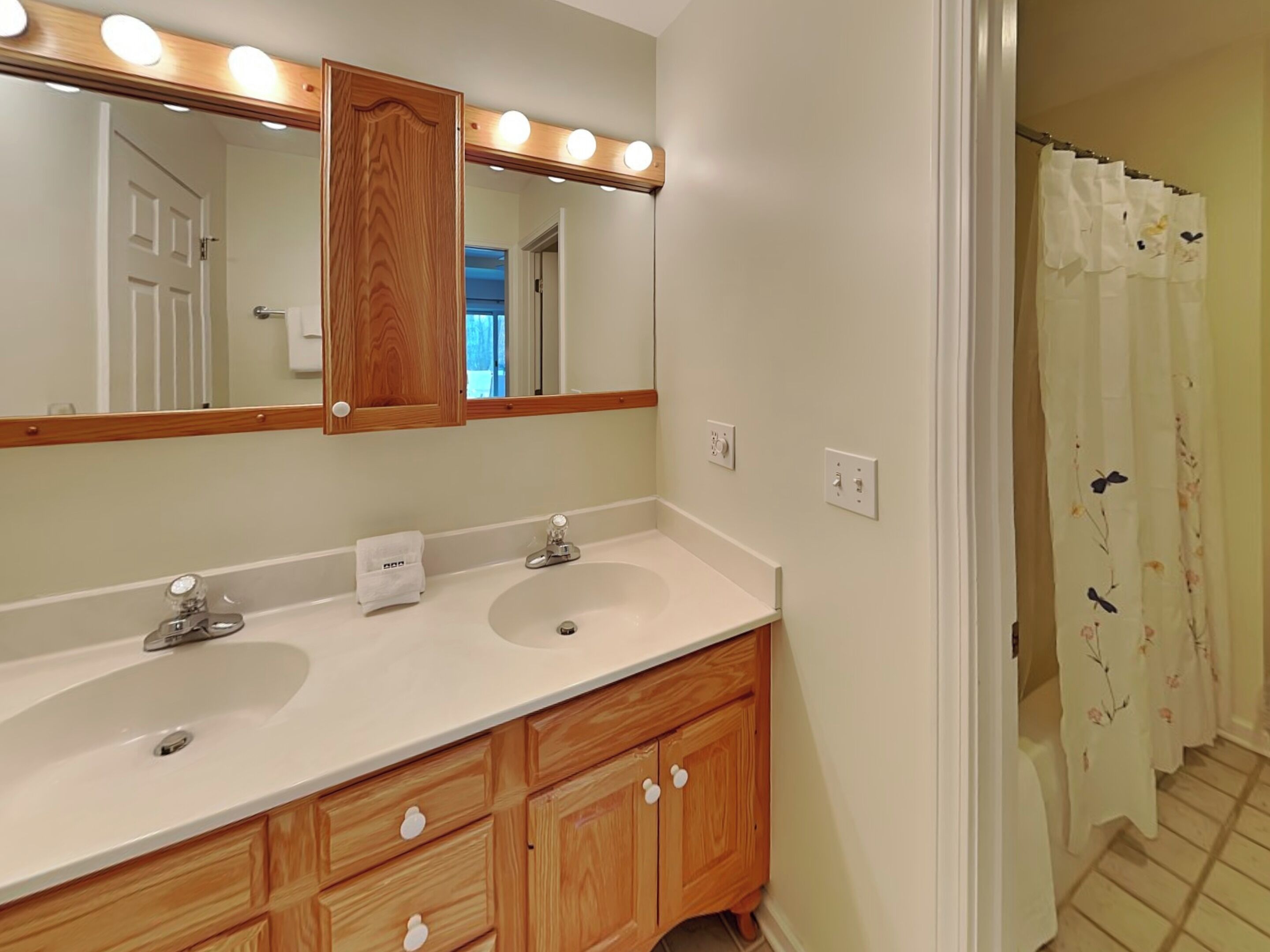 A bathroom with a double sink vanity, wooden cabinets, a large mirror with lights, and a shower curtain with a floral pattern.