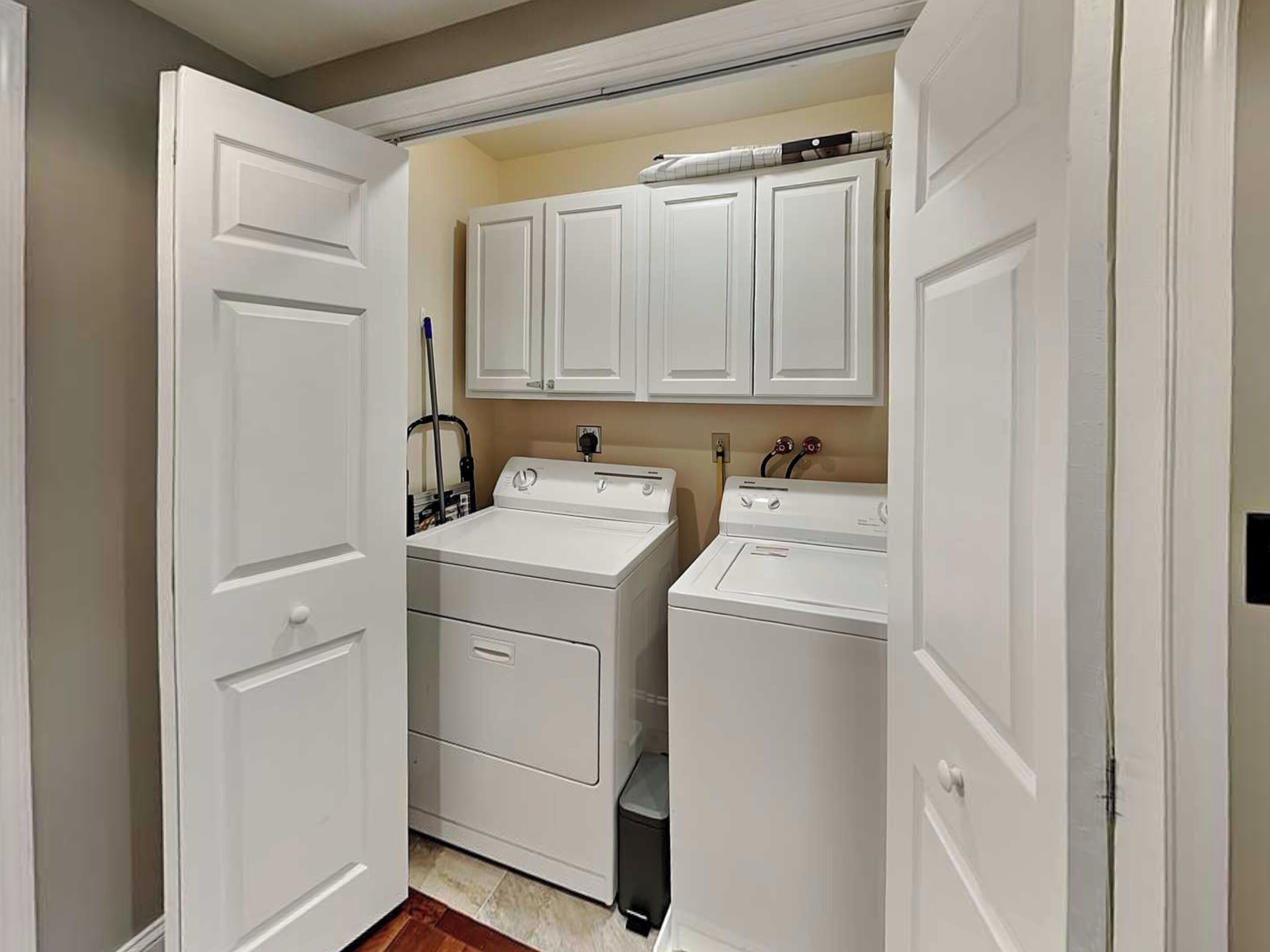A laundry room with two stacked washers and dryers, white cabinets above, and a mop leaning against the wall.