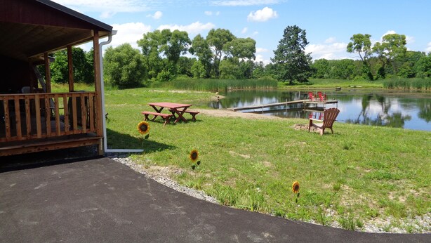 A lakeside picnic area with a wooden deck, picnic tables, and chairs, surrounded by lush greenery and trees.