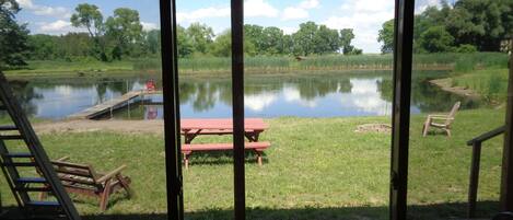 A view from inside a building looking out to a grassy area with a pond, a picnic table, and a wooden deck.