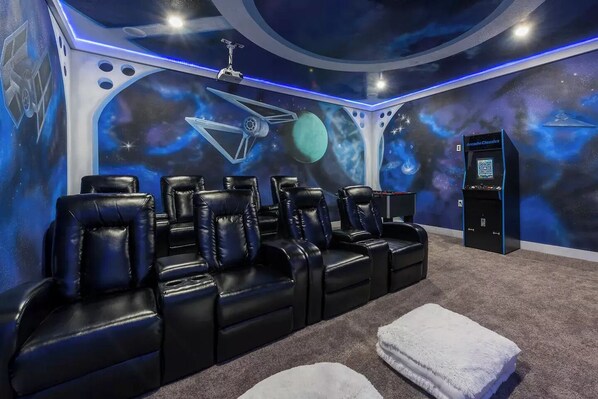 Una habitación con sillones reclinables de cuero negro, un proyector montado en el techo y un mural en la pared que representa escenas espaciales.