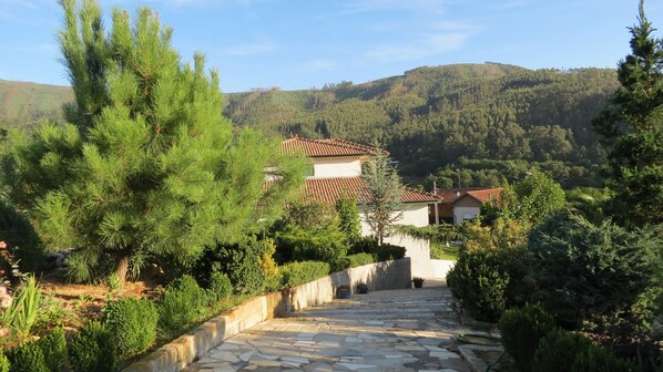 Un chemin de pierres mène à une maison dont le toit est recouvert de tuiles, et qui est entourée de verdure, avec des collines en arrière-plan.