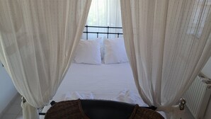 Un letto con lenzuola bianche e un baldacchino, un termosifone e una finestra con tende leggere.