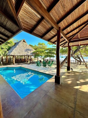 A pool area with a clear blue pool, wooden pergolas, and a thatched-roof structure.