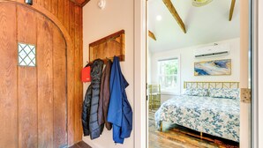Eine Holztür mit einem Kleiderhaken, an dem mehrere Jacken hängen, ein Bett mit einer blumigen Tagesdecke und eine Klimaanlage an der Wand.