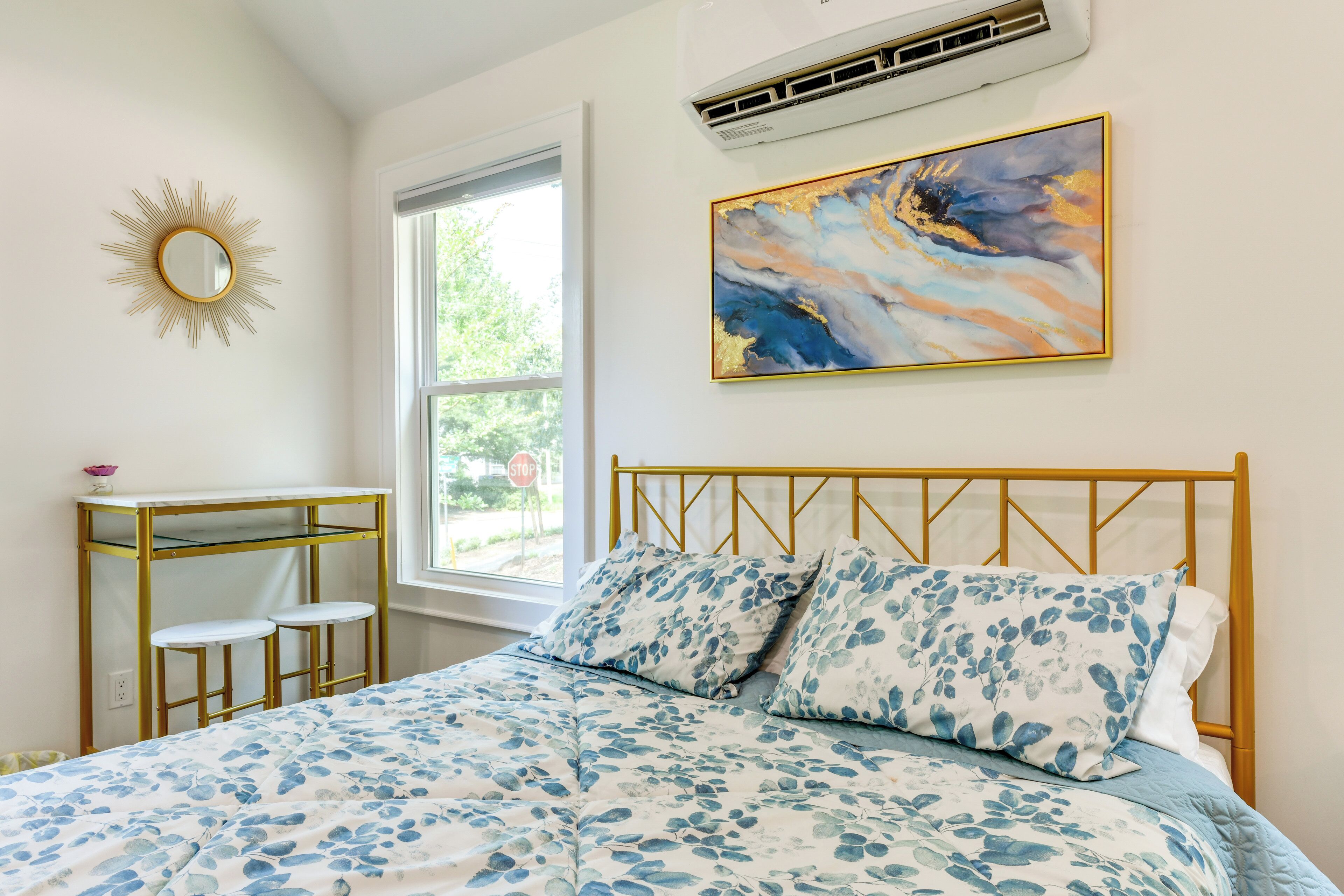A bedroom with a bed, a window, a wall-mounted air conditioner, a painting, and a sunburst mirror.