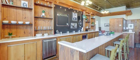 Una cocina moderna con armarios de madera, encimera blanca y taburetes de bar. Se ven estantes con vajilla y una pared de pizarra con un letrero de bienvenida.