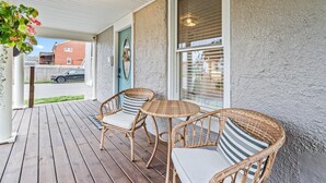 Varanda coberta com cadeiras de vime e uma pequena mesa, uma porta com espelho decorativo, e vista para a rua com um carro e uma casa ao fundo.