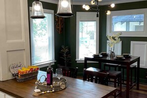 Un coin repas avec une table en bois, des chaises et un lustre. Il y a des fenêtres avec des stores et une décoration murale.