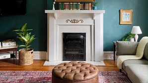 Ein Wohnzimmer mit Kamin, einem aufgehängten Gemälde, einem Bücherregal, einem Fernseher, einer Couch, einem Hocker und einem gemusterten Teppich.