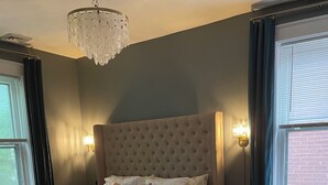 Ein Schlafzimmer mit einem großen Bett, einer Chandelieber, zwei Nachttischlampen, einem Teppich und einem Fenster mit Vorhängen.