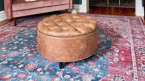 Ein Wohnzimmer mit einem rosafarbenen Sofa, einem runden Hocker, einem gemusterten Teppich und einem Essbereich mit Holzkohlen und einem Tisch.