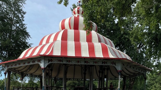 Ein Picknick mit kariertem Tischlein, Speisen und Getränken auf einer Wiese, im Hintergrund ein Pavillon.