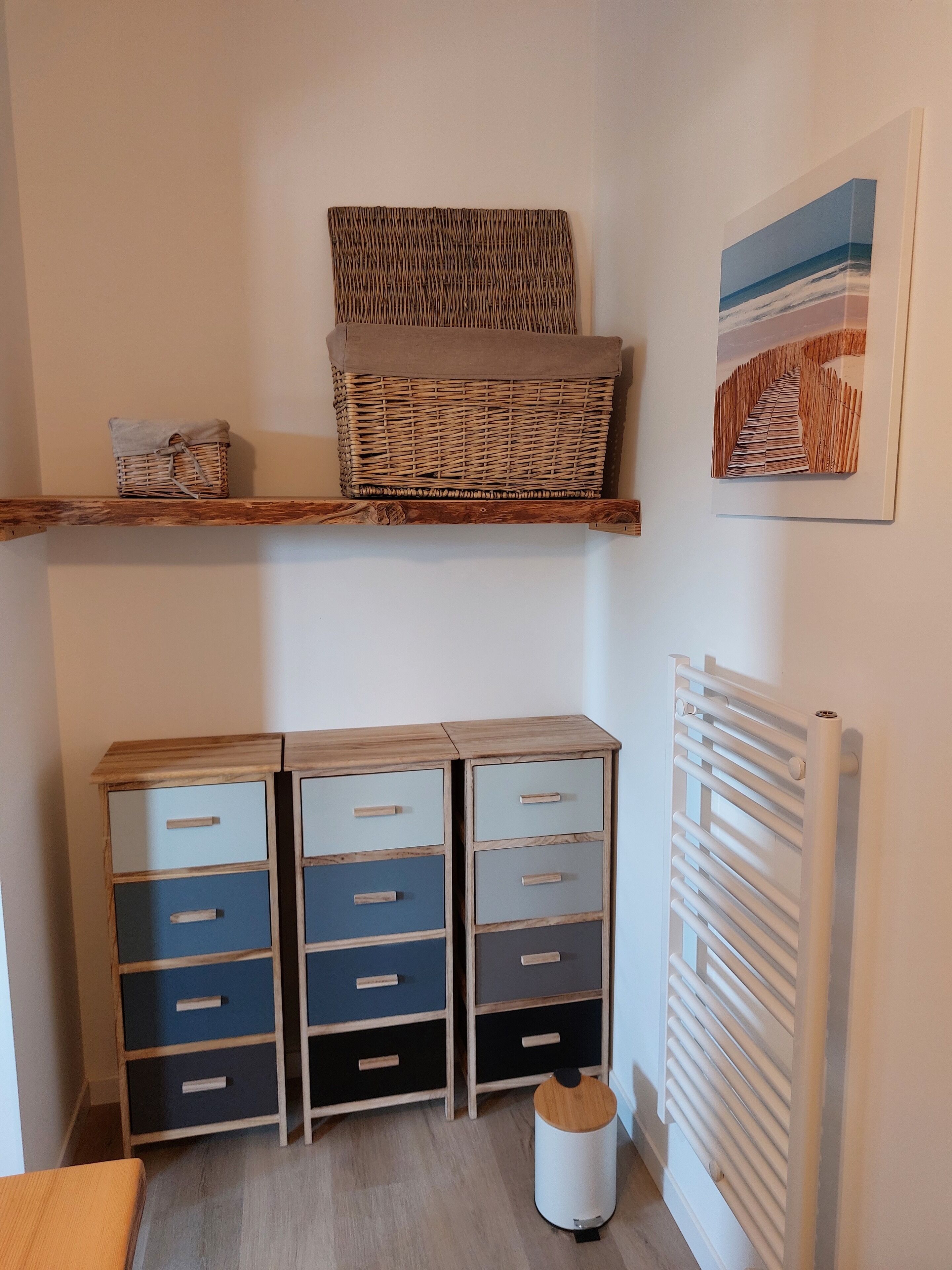A room with a wooden cabinet with drawers, a wicker basket on a shelf, a radiator, and a framed picture on the wall.