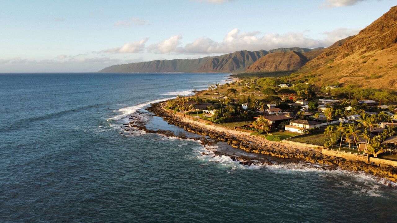 A coastal town with houses along the shoreline, a rocky breakwater, and mountains in the background.
