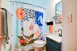 Un baño con cortina de ducha estampada, inodoro blanco, lavabo con grifo negro y espejo sobre el lavabo.