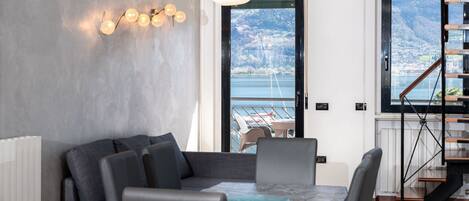 Ein modernes Wohnzimmer mit einer Couch, einem Esstisch und Stühlen. Eine Treppe führt zu einem Balkon mit Blick auf das Meer und die Berge.