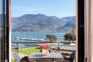 A balcony with a table and chairs, a vase of flowers, and a view of a marina and mountains. - magnificent apartment with terrace overlooking the lake. (Lovere)