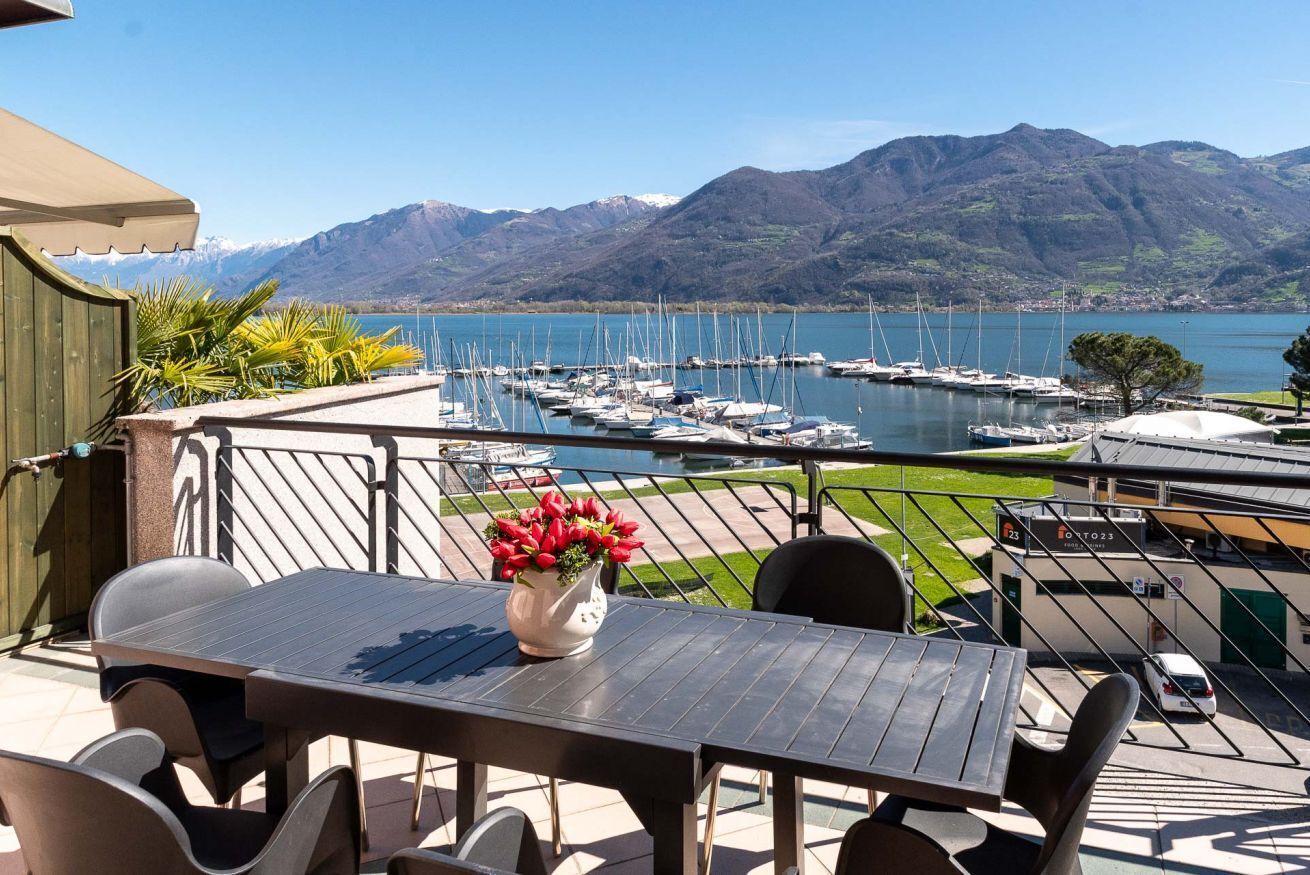 Une terrasse avec une table et des chaises, donnant sur une marina où sont amarrés des bateaux, avec des montagnes en arrière-plan.