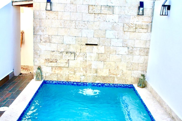 Una piscina pequeña y rectangular con borde de azulejos, rodeada de muros de piedra y dos luces de pared tipo farol.